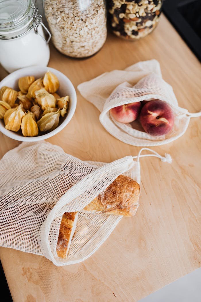 Close-up of reusable mesh bags with bread and fruit on a wooden kitchen surface.