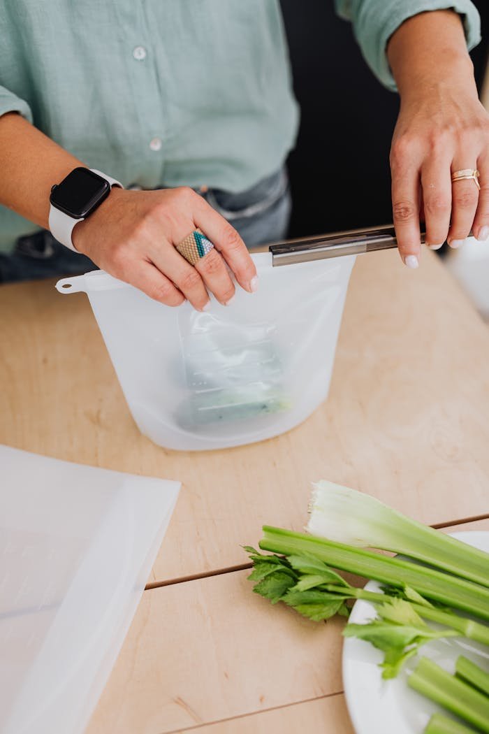 Hands sealing a reusable silicone bag on a kitchen table with fresh celery.