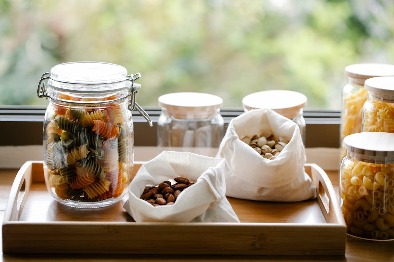 Glass jar with rotini pasta placed on wooden tray near ECO friendly bags with almonds and pistachios in light room near window on blurred background